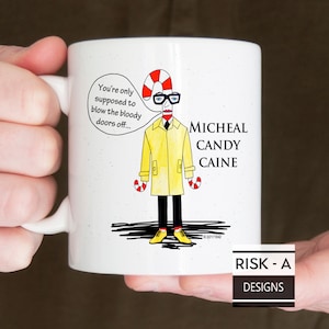 May include: White ceramic mug with a cartoon illustration of a person with candy cane arms and legs wearing a yellow raincoat and glasses. The text on the mug reads "You're only supposed to blow the bloody doors off... Micheal Candy Caine".
