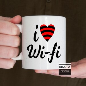 May include: White ceramic coffee mug with a black and red heart with a wifi symbol inside. The text "i love Wi-Fi" is written in black cursive lettering. The mug is held by a hand. The text "RISK - A DESIGNS" is printed in black on a white rectangle at the bottom of the mug.