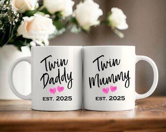 May include: Two white ceramic mugs with black text that reads "Twin Daddy" and "Twin Mummy" with pink heart graphics and the year "EST. 2025". The mugs are on a wooden surface with a blurred floral background.