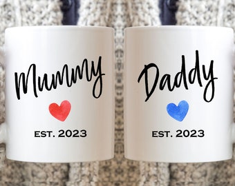May include: Two white ceramic mugs with black lettering. The mug on the left says "Mummy EST. 2023" with a red watercolor heart. The mug on the right says "Daddy EST. 2023" with a blue watercolor heart.