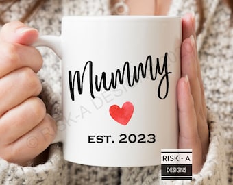 May include: White ceramic mug with black text that reads "Mummy" and a red watercolor heart. Below the text is "EST. 2023".