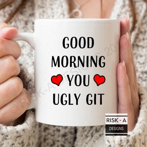 Puede incluir: Taza de cerámica blanca con texto negro que dice "Good Morning You Ugly Git" con dos corazones rojos. La taza está siendo sostenida por las manos de una persona.