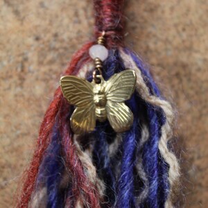May include: A close-up of a colourful yarn hair extension with a gold butterfly charm and a white bead.