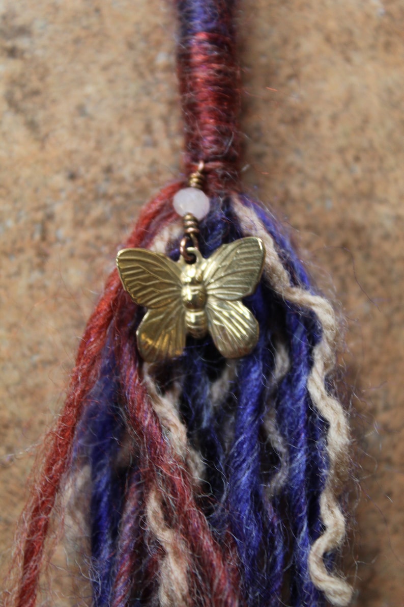 May include: A close-up of a colourful yarn hair extension with a gold butterfly charm and a white bead.