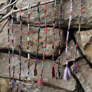 May include: A collection of colourful, braided hair extensions hanging from a branch. The extensions are made of various materials, including yarn, thread, and fabric. They are all different lengths and have different patterns.