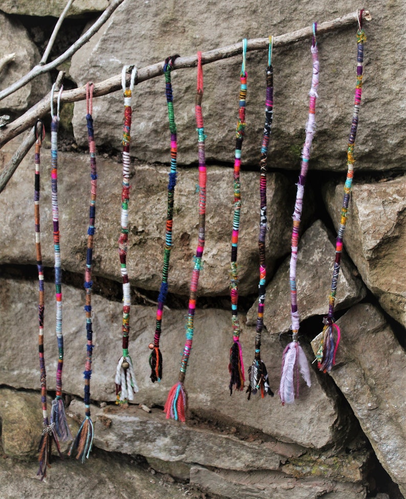 May include: A collection of colourful, braided hair extensions hanging from a branch. The extensions are made of various materials, including yarn, thread, and fabric. They are all different lengths and have different patterns.