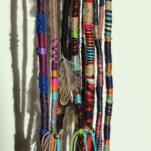 May include: A set of colourful yarn hair extensions with feathers and beads. The extensions are made of various colours, including purple, blue, green, red, orange, yellow, and brown. The extensions are tied together with a small metal ring.