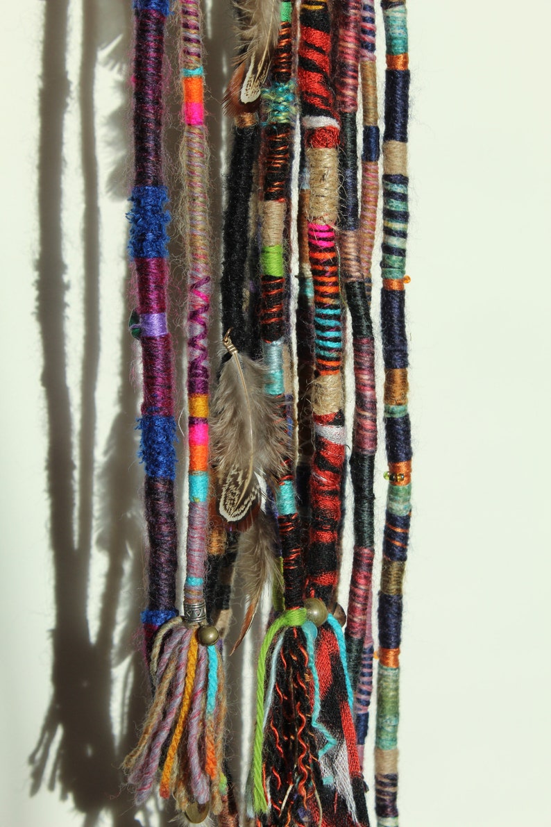 May include: A set of colourful yarn hair extensions with feathers and beads. The extensions are made of various colours, including purple, blue, green, red, orange, yellow, and brown. The extensions are tied together with a small metal ring.