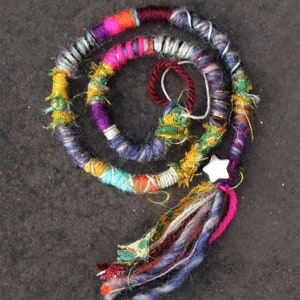 May include: A colourful, multi-stranded hair wrap with a white star charm. The wrap is made of various yarns in shades of pink, purple, blue, green, yellow, and white. The wrap is tied with a tassel at the end.