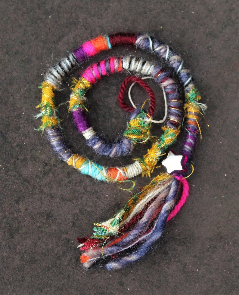 May include: A colourful, multi-stranded hair wrap with a white star charm. The wrap is made of various yarns in shades of pink, purple, blue, green, yellow, and white. The wrap is tied with a tassel at the end.