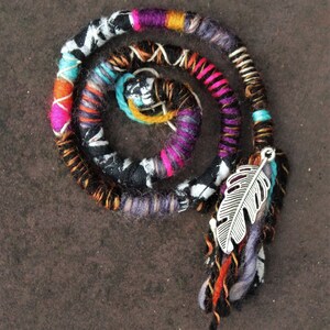 May include: A colourful hair wrap with a silver feather charm. The wrap is made of multiple strands of yarn in various colours, including black, white, pink, purple, orange, and turquoise. The feather charm is attached to the wrap with a silver ring.