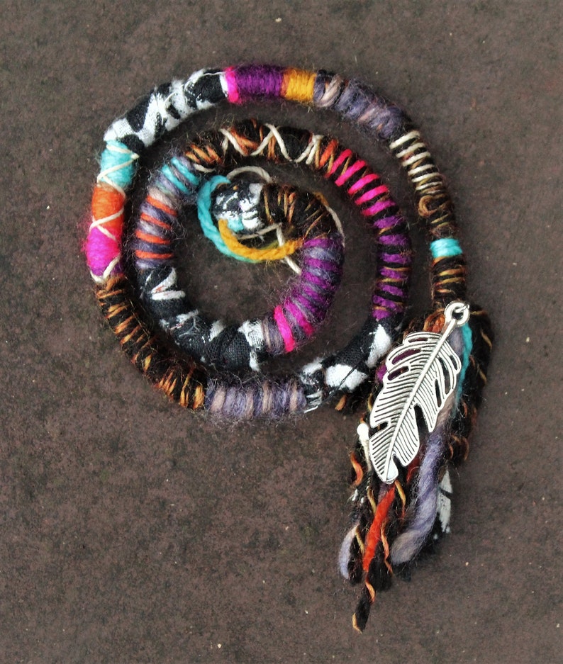 May include: A colourful hair wrap with a silver feather charm. The wrap is made of multiple strands of yarn in various colours, including black, white, pink, purple, orange, and turquoise. The feather charm is attached to the wrap with a silver ring.