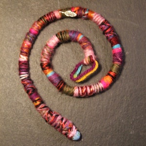 May include: A colourful, multi-stranded yarn hair wrap with a silver leaf charm. The wrap is coiled into a circle and has a variety of colours, including pink, red, orange, yellow, green, blue, and purple.