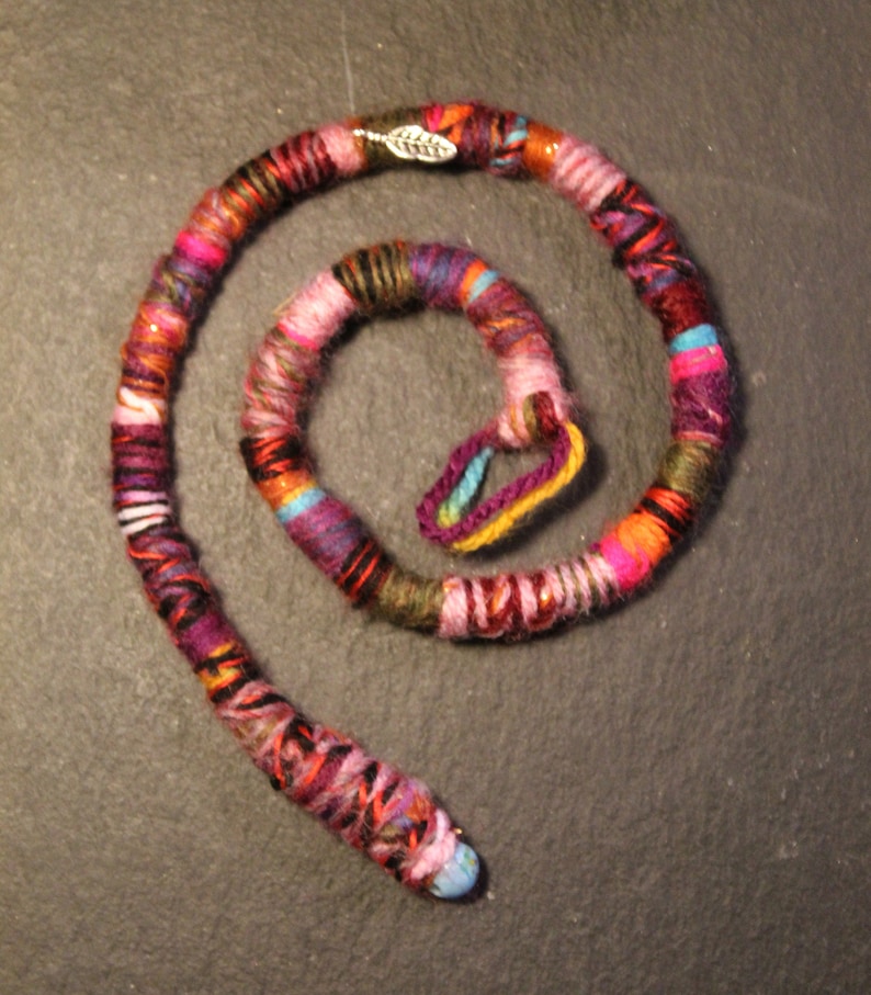 May include: A colourful, multi-stranded yarn hair wrap with a silver leaf charm. The wrap is coiled into a circle and has a variety of colours, including pink, red, orange, yellow, green, blue, and purple.