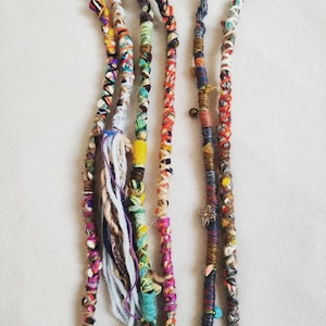 May include: Five colourful crocheted hair extensions with beads and charms. The extensions are made of various colours of yarn, including blue, green, red, yellow, and white.