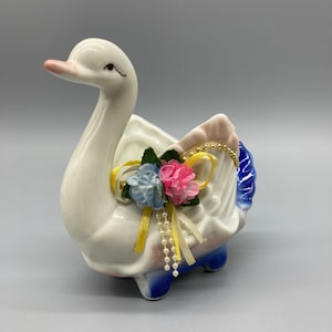 May include: A white ceramic swan planter with a pink beak and blue accents. The swan is decorated with artificial pink and blue flowers, gold ribbon, and a pearl-like strand. The planter has a decorative design and is suitable for small plants or floral arrangements.