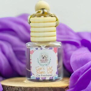 May include: A clear glass car air freshener with a wooden top and a label that reads "Sun's Out Buns Out Car Freshener". The air freshener sits on a small wooden slice, with a purple fabric draped around it.