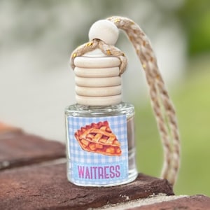 May include: A small, clear glass bottle with a wooden cap and a woven cord. The bottle has a label with a slice of pie on a blue gingham background and the word "Waitress". The bottle is sitting on a red brick surface.