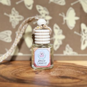 Peut inclure: Un petit désodorisant pour voiture en verre avec un bouchon en bois et une étiquette Vanilla Cinnamon. L'étiquette présente un motif floral et le texte "Vanilla Cinnamon Car Freshener". Le désodorisant est suspendu à une ficelle beige et jaune.
