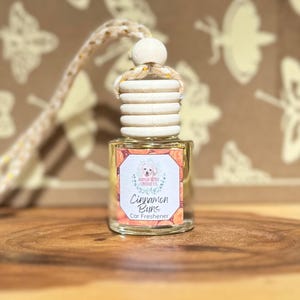 May include: A clear glass car air freshener bottle with a wooden cap and a string for hanging. The label reads "Cinnamon Buns Car Freshener" with a dog illustration. The background is a brown surface with butterfly patterns.