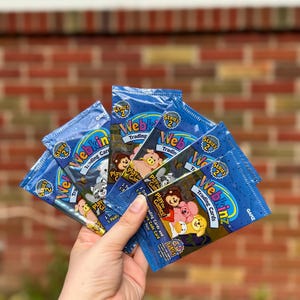 May include: Five unopened packs of Webkinz Series 2 Trading Cards. Each pack features a blue background with a cartoon animal and the text "Webkinz Trading Cards" in white. The packs are held in a hand.