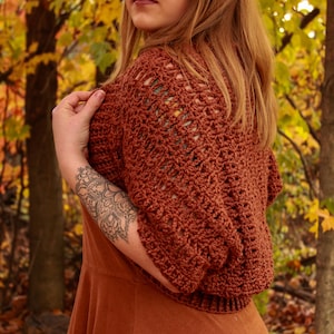 Puede incluir: Un bolero de ganchillo calado de color óxido. La prenda de manga corta tiene un patrón texturizado y de encaje. La modelo lleva un vestido color óxido. El fondo presenta follaje otoñal.