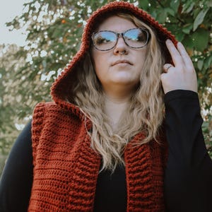 Pode incluir: Um close-up de uma mulher vestindo um colete de crochê cor ferrugem com capuz. O colete tem um decote em V profundo e é sem mangas. A mulher está vestindo uma camisa preta de manga comprida por baixo do colete. Ela está usando óculos e tem cabelo loiro.