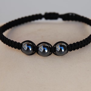 May include: A black macrame bracelet with three polished, dark grey beads. The bracelet is adjustable with sliding knots and is displayed on a beige cushion. The beads are approximately 1.3 cm in diameter.