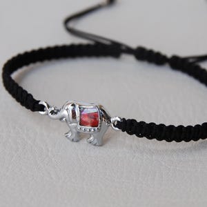 Può includere: Bracciale intrecciato nero con un ciondolo a forma di elefante argentato con un design rosso e bianco. Il bracciale ha cordini neri regolabili. Un gioiello semplice ma elegante.