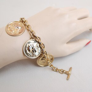 Puede incluir: Un brazalete dorado con una cadena y múltiples dijes redondos en forma de moneda. Los dijes tienen un diseño en relieve de un perfil de rostro humano.