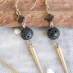 May include: A pair of dangle earrings with gold-colored hooks. Each earring features a small black cross, a textured black sphere, and a gold-colored spike. The earrings are displayed on a wooden surface.