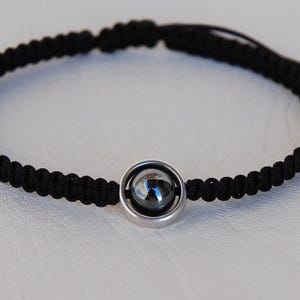 May include: Black braided bracelet with a central silver-toned bead. The bracelet is adjustable with sliding knots and features a small, dark-colored stone in a silver setting. The bracelet is displayed on a white surface.