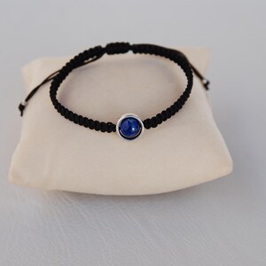 Op de afbeelding: Zwart macram&eacute; armband met een donkerblauwe steen in zilver gezet. De armband wordt gepresenteerd op een klein, cr&egrave;mekleurig kussen.
