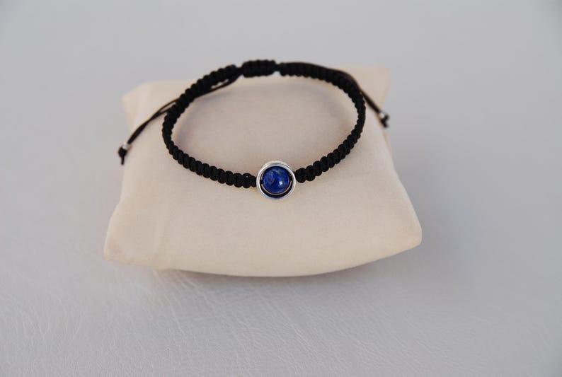 Op de afbeelding: Zwart macram&eacute; armband met een donkerblauwe steen in zilver gezet. De armband wordt gepresenteerd op een klein, cr&egrave;mekleurig kussen.
