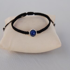 Op de afbeelding: Zwart macram&eacute; armband met een zilverkleurige zetting en een ronde, diepblauwe steen. De armband wordt gepresenteerd op een cr&egrave;mekleurig kussen. Het verstelbare ontwerp zorgt voor een comfortabele pasvorm.