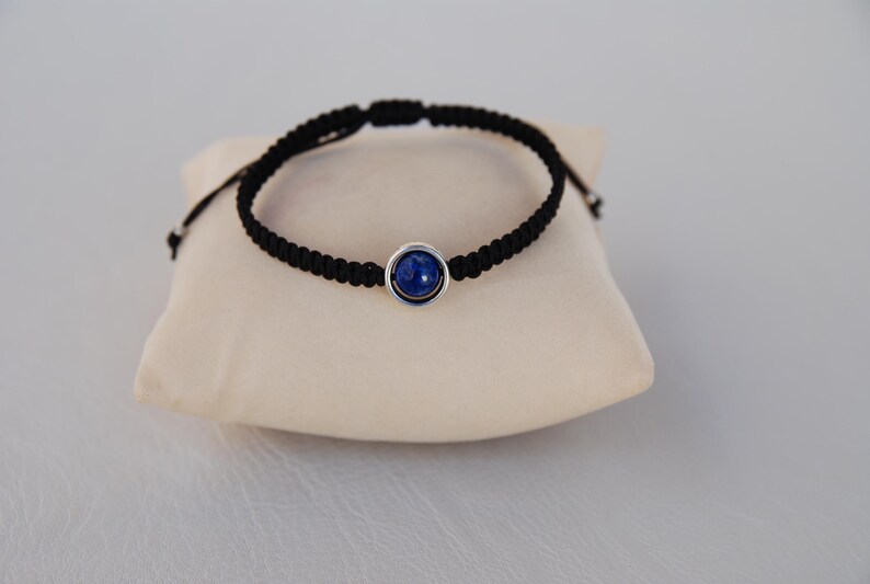 Op de afbeelding: Zwart macram&eacute; armband met een zilverkleurige zetting en een ronde, diepblauwe steen. De armband wordt gepresenteerd op een cr&egrave;mekleurig kussen. Het verstelbare ontwerp zorgt voor een comfortabele pasvorm.