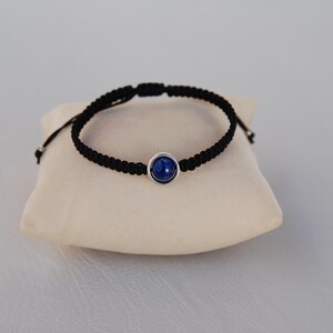 Op de afbeelding: Een zwart macram&eacute; armband met een ronde, diepblauwe steen in een zilverkleurige bezel. De armband wordt getoond op een klein, cr&egrave;mekleurig kussen. De verstelbare armband is een stijlvol accessoire.