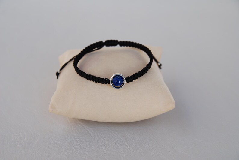 Op de afbeelding: Een zwart macram&eacute; armband met een ronde, diepblauwe steen in een zilverkleurige bezel. De armband wordt getoond op een klein, cr&egrave;mekleurig kussen. De verstelbare armband is een stijlvol accessoire.