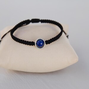 Op de afbeelding: Een zwart macram&eacute; armband met een centrale blauwe edelsteen gezet in een zilverkleurige bezel. De armband wordt gepresenteerd op een cr&egrave;mekleurig kussen, wat het ontwerp van de sieraden benadrukt. Het verstelbare koord zorgt voor een comfortabele pasvorm.