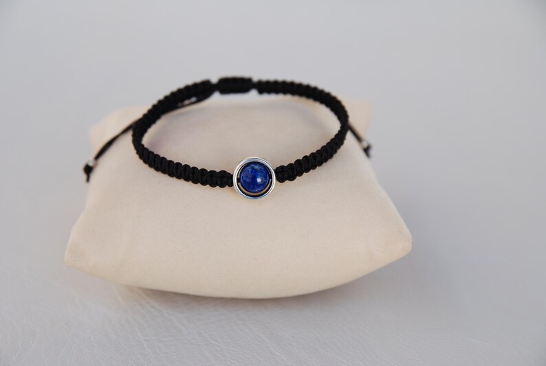 Op de afbeelding: Een zwart macram&eacute; armband met een centrale blauwe edelsteen gezet in een zilverkleurige bezel. De armband wordt gepresenteerd op een cr&egrave;mekleurig kussen, wat het ontwerp van de sieraden benadrukt. Het verstelbare koord zorgt voor een comfortabele pasvorm.