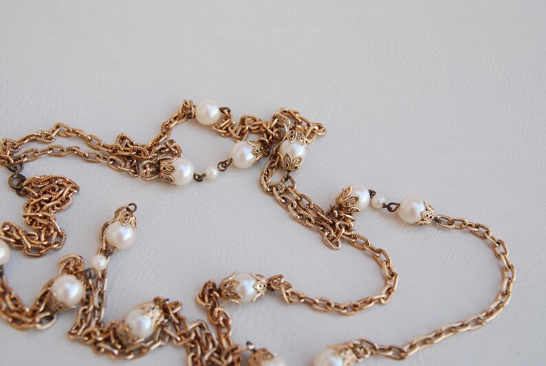 Puede incluir: Un collar de cadena dorada con cuentas de perlas blancas. La cadena est&aacute; hecha de m&uacute;ltiples hebras y tiene un dise&ntilde;o delicado e intrincado.