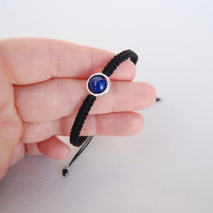 Op de afbeelding: Zwart gevlochten armband met een ronde, diepblauwe edelsteen in een zilverkleurige zetting. De armband is verstelbaar met zwarte koorden en een schuifknoopsluiting. Een eenvoudig, elegant sieraad.