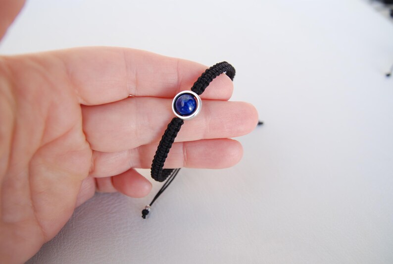 Op de afbeelding: Zwart gevlochten armband met een ronde, diepblauwe edelsteen in een zilverkleurige zetting. De armband is verstelbaar met zwarte koorden en een schuifknoopsluiting. Een eenvoudig, elegant sieraad.