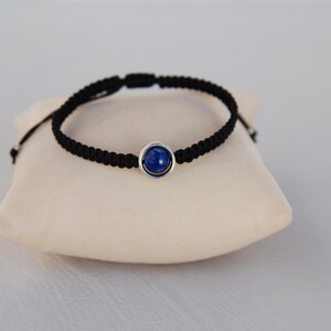 Op de afbeelding: Zwart gevlochten armband met een zilverkleurige zetting met een diepblauwe edelsteen. De armband wordt gepresenteerd op een beige kussen tegen een lichtgrijze achtergrond. Eenvoudig, elegant ontwerp.