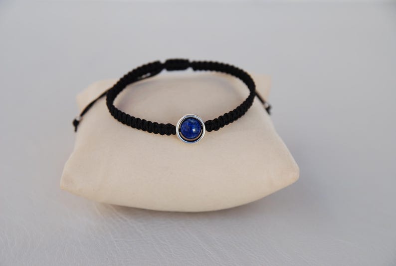 Op de afbeelding: Zwart gevlochten armband met een zilverkleurige zetting met een diepblauwe edelsteen. De armband wordt gepresenteerd op een beige kussen tegen een lichtgrijze achtergrond. Eenvoudig, elegant ontwerp.