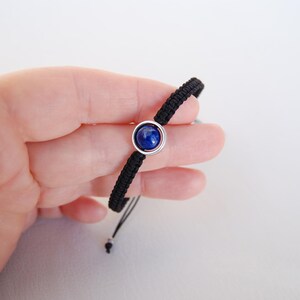 Op de afbeelding: Zwart macram&eacute; armband met een centrale, ronde, diepblauwe edelsteen gezet in een zilverkleurige bezel. De armband wordt tegen een witte achtergrond gehouden.