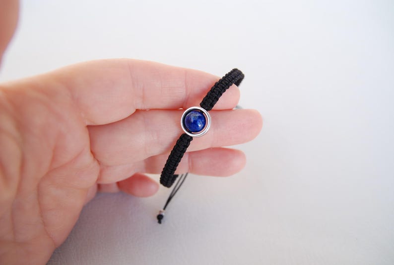 Op de afbeelding: Zwart macram&eacute; armband met een centrale, ronde, diepblauwe edelsteen gezet in een zilverkleurige bezel. De armband wordt tegen een witte achtergrond gehouden.