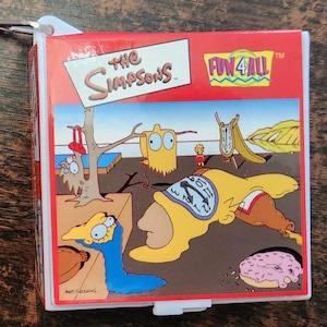 Może przedstawiać: Czerwono-biały brelok do kluczy z kreskówkowym obrazkiem postaci z Simpsonów. Brelok ma metalowy pierścień i łańcuszek. Na breloku nadrukowany jest tekst "The Simpsons" i "Fun 4 All".