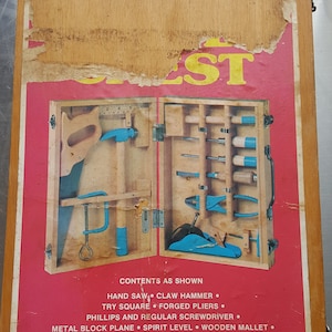 May include: A red and blue wooden toy tool chest with a variety of tools, including a hand saw, claw hammer, try square, forged pliers, Phillips and regular screwdriver, metal block plane, spirit level, wooden mallet, chisel, "C" clamp, and gimlet. The text "CONTENTS AS SHOWN" is printed on the front of the chest.