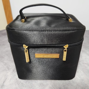 Könnte beinhalten: Schwarze Kosmetiktasche aus Satin mit oberem Griff und goldfarbener Hardware. Die Tasche hat einen Reißverschluss und ein rechteckiges, goldfarbenes Etikett mit der Aufschrift "GIORGIO BEVERLY HILLS".